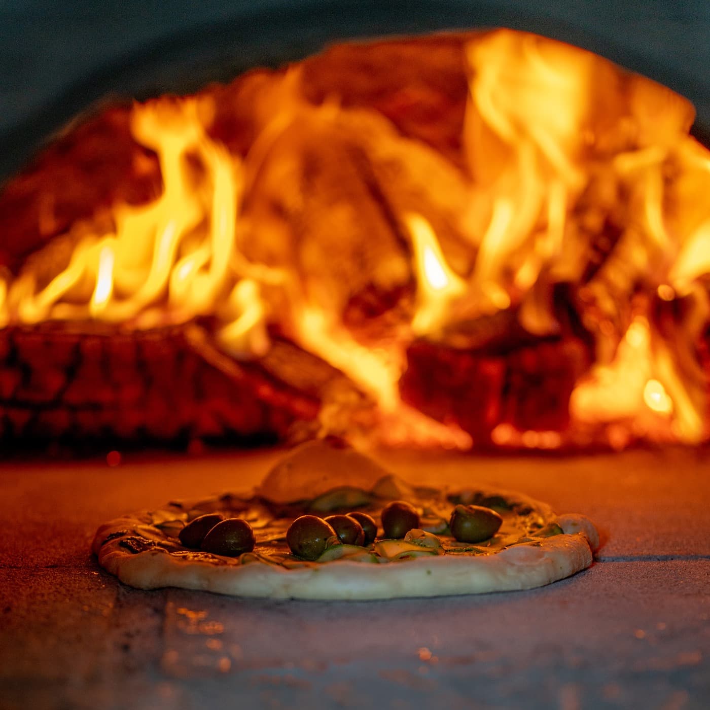 Pizza assando no lastro do forno a lenha, com as chamas altas ao fundo